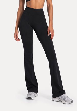 Mujer con pantalones negros de talle alto, ajustados y acampanados, y zapatillas blancas, de pie con las manos relajadas a los lados sobre un fondo blanco.