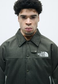 Olíva zöld halászmintás dzseki mandarin gallérral, gombos záródással és fehér logóval a bal mellen.