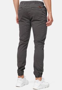 Man draagt donkergrijze joggingbroek met elastische boorden en witte sneakers, gezien van achteren tegen een effen achtergrond.