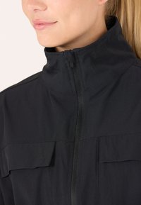 Black jacket with high collar, front zipper, and two chest pockets. Material appears smooth and lightweight with a structured design.