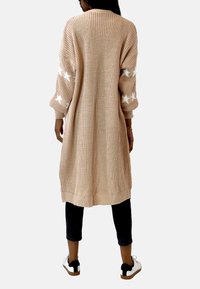Lång, stickad cardigan i beige med vita stjärnmönster på ärmarna. Har en avslappnad passform, ribbade ärmslut och en strukturerad yta.