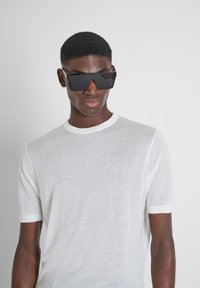 Camisa blanca de manga corta hecha de un material ligero y transparente, combinada con gafas de sol negras angulares, sobre un fondo neutro.