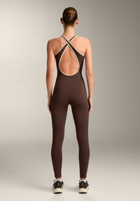 Woman standing facing away wearing a fitted brown jumpsuit with crisscross open back and beige trim, paired with black and white sneakers.