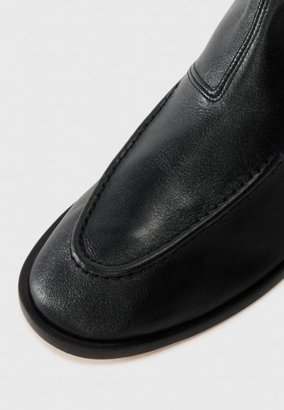 Chaussure en cuir noir avec un bout arrondi, texture lisse, détails brodés le long des bords, et un design élégant sans matériel visible.