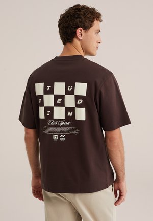 Mann trägt dunkelbraunes T-Shirt mit weißem Schachbrettmuster und stilisiertem Schriftzug „TÜZEDIN Club Spirit“ auf dem Rücken, steht seitlich.