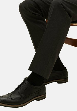 Scarpe brogue in pelle nera con dettagli perforati, abbinate a pantaloni grigi scuri su misura, sedute su una sedia di legno.