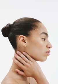 Aro de oro, collar de cadenas de oro en capas y anillo texturizado en los dedos. Piel suave y cabello peinado en un moño bajo.