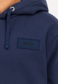 Nærbilde av en marinblå hettegenser med snor og Alpha Industries-logo på brystet.