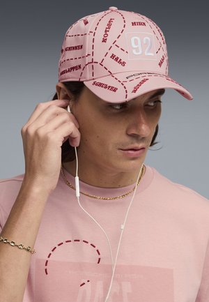 Persona joven que lleva una gorra rosa con términos anatómicos rojos y auriculares blancos, tocándose una oreja, vestida con una camiseta rosa y cadena y pulsera doradas.