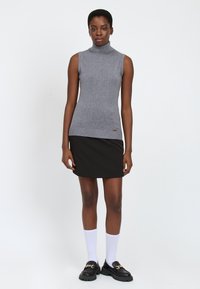 Top grigio senza maniche a collo alto in tessuto a maglia, abbinato a una mini gonna nera. La modella indossa calze bianche fino al ginocchio e mocassini neri.