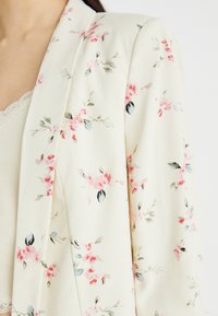 Blazer crème clair avec des motifs floraux roses et verts. Tissu doux et lisse, design à ouverture frontale et col châle.