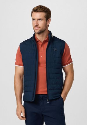 Gilet matelassé bleu marine avec col haut, fermeture éclair à l'avant et détail de logo, porté sur un polo corail. Texture lisse et design léger.