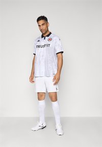 Maillot de football blanc et gris avec un col noir, incluant un logo et le texte "FAIS-TOI CONFIANCE". Porté avec un short blanc et des chaussettes, complété par des crampons noirs et blancs.