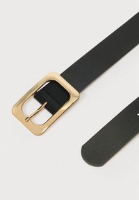 Mustast nahast vöö, millel on ristküliku kujuline kuldne pandl ja sile pind. Vöö ots on ümar ning kujundus on minimalistlik.