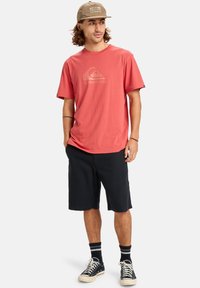 Korálové bavlněné tričko s logem Quiksilver, kombinované s černými šortkami a černými teniskami. K tomu se dokonale hodí khaki kšiltovka, což dotváří ležérní vzhled.