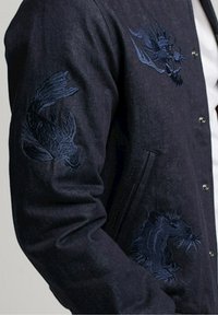 Veste marine ornée de motifs brodés en bleu foncé représentant un poisson et un dragon sur les manches, avec une fermeture à boutons-pression et une texture lisse.