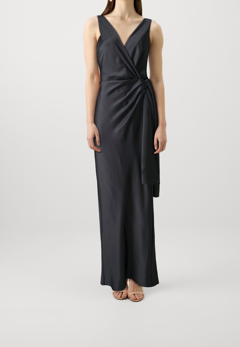 Kobieta ubrana w czarną satynową sukienkę kopertową bez rękawów z detalem węzła po bokach, stoi na tle jasnego, gładkiego tła.