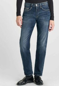 Jeans de mezclilla azul oscuro con corte recto, cinco bolsillos y costuras en contraste. Llevados con mocasines negros y una blusa de manga larga.