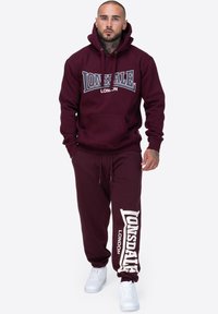 Burgunderfarbene Hoodie- und Jogginganzug-Sets aus Baumwolle. Mit dem Lonsdale-Logo in kontrastierendem Weiß, Kapuze und Fronttasche. Weiße Sneakers.