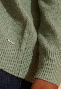 Miętowo-zielony sweter dziany z prążkowanymi mankietami. Posiada małą metalową metkę z grawerunkiem. Teksturowany materiał i miękka faktura.