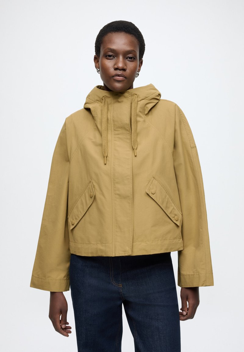 Rövidített szabású, mustársárga kapucnis dzseki, két nagy oldalzsebbel és egyenes szabással. Simább, könnyű anyagból készült.