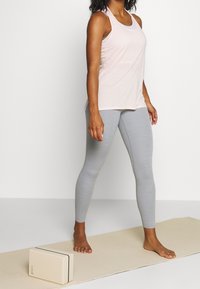 Top in rosa chiaro senza maniche e leggings grigi chiari, in piedi su un tappetino da yoga beige con un blocco da yoga vicino.