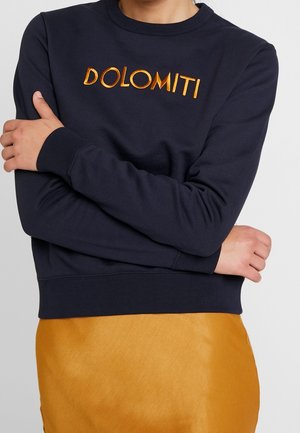 Osoba nosząca granatową bluzę z haftem "DOLOMITI" w kolorze pomarańczowym oraz musztardowożółtą spódnicę, z założonymi na piersi skrzyżowanymi rękami.