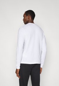 Maglietta bianca a maniche lunghe in morbido cotone con scollo rotondo, orlo dritto e polsini a costine, indossata con pantaloni grigio scuro.