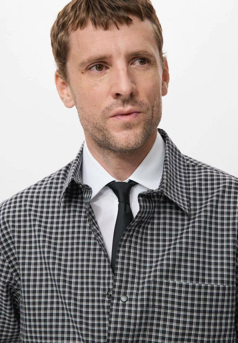 Black and white checkered shirt with a contrasting white collar, featuring a front pocket and button closure, worn over a black tie.