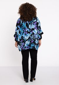 Florale kimono in zwart met blauwe en paarse bladvormen. Losse pasvorm met brede mouwen, gecombineerd met zwarte broek en blokhakken schoenen.