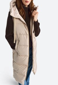 Beige puffer vest dengan penutup kepala, depan resleting, dan saku samping. Memiliki tekstur quilted dan tali beige. Dikenakan di atas sweter cokelat.