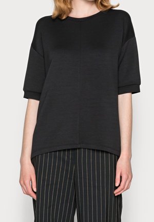 Femme portant un haut noir ample à manches courtes associé à un pantalon noir à fines rayures blanches verticales.
