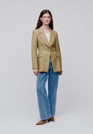 Kvinde med krøllet hår iført lysebrun blazer, hvid top, blå jeans med vide ben og brune spidse hæle, stående foran en ensfarvet baggrund.