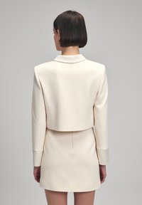 Blazer court crème à manches longues et col cranté, associé à une mini-robe ajustée assortie. Tissu lisse et structuré.