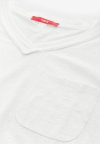 White cotton T-shirt with a V-neck, featuring a left chest pocket and a red brand label at the neckline. Soft, plain texture.
