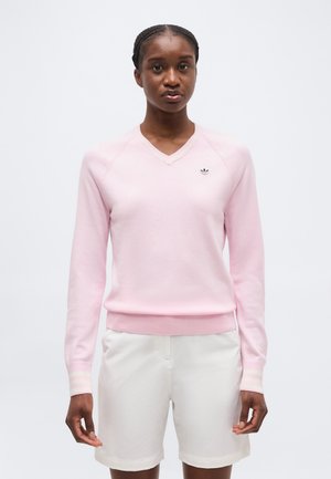 Γυναίκα που φοράει πουλόβερ μακρυμάνικο ανοιχτό ροζ με V λαιμόκοψη και μικρό λογότυπο, συνδυασμένο με λευκά σορτς μέχρι τα γόνατα, στέκεται μπροστά σε μονόχρωμο φόντο.