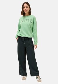 Felpa verde menta con testo "GIOIOSO", abbinata a pantaloni neri larghi e sneakers, con lacci a contrasto e una vestibilità comoda.