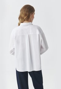 White button-up shirt with a collar, dropped shoulders, and a back pleat. Oversized fit with smooth texture, paired with dark pants.