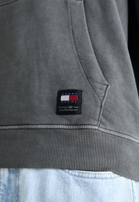 Harmaa collegepaita, jossa on resoroitu helma ja tasku. Etupuolella suorakaiteen muotoinen merkki, jossa mustaa, valkoista ja punaista, ja teksti "TOMMY JEANS."