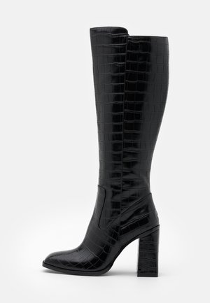 Botte noire montante jusqu'au genou avec motif crocodile, bout pointu et talon haut épais sur fond blanc.