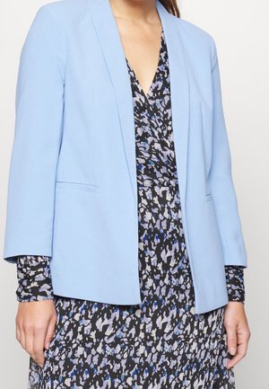Vrouw draagt een lichtblauw blazer over een zwarte, blauwe en grijze abstracte patroonjurk met lange mouwen.