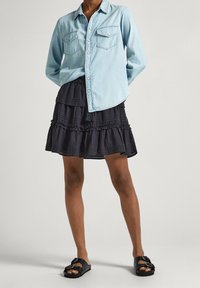 Osoba v světle modré košili s knoflíky z džínsoviny, černé sukni se sklady a černých sandálech na přezku, stojící na jednobarevném pozadí.