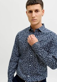 Noor mees lühikeste pruunide juustega, kellel on seljas tumelõheroheline lillemustriga nööbitav särk, kohandamas kraed ühtlase heleda tausta vastu.