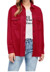 Kvinde iført rød skjorte med knapper og klaplommer over hvid grafisk t-shirt og lyseblå mønstrede jeans, vist fra halsen til hoften.