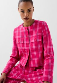 La chaqueta de traje a cuadros rosa presenta un escote redondo, dos bolsillos frontales y cierre de botones, elaborada con un tejido texturizado con líneas blancas contrastantes.