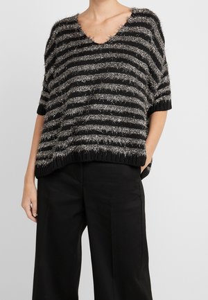 Femme portant un pull court à manches courtes duveteux rayé noir et argent avec un pantalon noir à jambes larges, une main dans la poche, sur un fond uni.