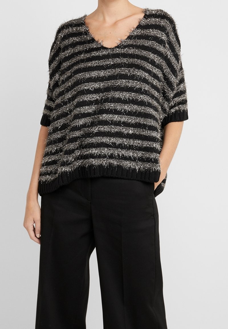 Femme portant un pull court à manches courtes duveteux rayé noir et argent avec un pantalon noir à jambes larges, une main dans la poche, sur un fond uni.