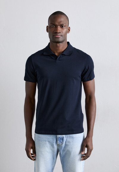 Polo shirt blu navy realizzato in morbido tessuto di cotone. Maniche corte, colletto classico e design aderente. Indossato con jeans azzurri chiari.
