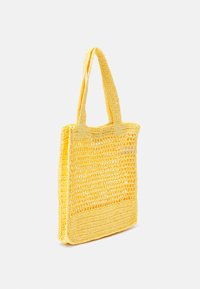 Sac fourre-tout en crochet jaune avec deux bretelles, présentant un design en mailles ouvertes et une base plate pour plus de stabilité.
