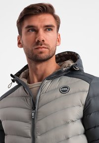 Vaatteiden suojakerroksena toimii harmaa ja musta toppatakki, jossa on vaakasuuntainen tikkauskuviointi, vetoketjukiinnitys, säädettävä huppu ja rintataskussa logo.